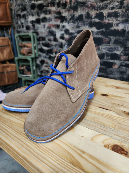 A pair of beige vellies with blue laces and sole detail, displayed on a wooden surface with a brick wall background.