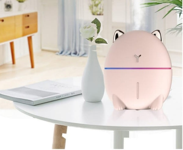 A pink, cat-shaped mini humidifier on a white table in a cozy room setting.